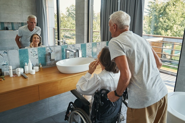 smiling elderly couple staring into bathroom mirror where countertop has been lowered for handicap accessibility; woman is in wheelchair and man is standing behind wheelchair