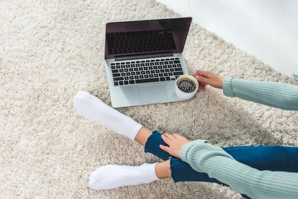 closeup of woman sitting on new carpet installation, showing her legs near a laptop on the floor, hands holding a cup of coffee