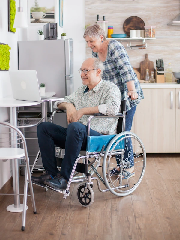 elderly couple in home kitchen looking at laptop on a table; man is in wheelchair and woman behind him pushing wheelchair closer to the table, modified with aging-in-place remodeling